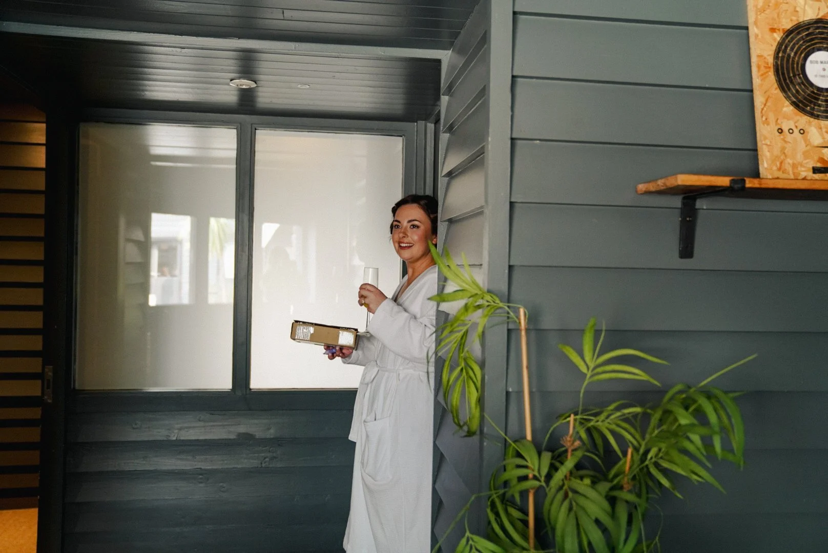 Woman in a white bathrobe holding a glass of champagne and a box, standing partly behind a wall with green plants, inside a modern room with large windows and dark gray walls.