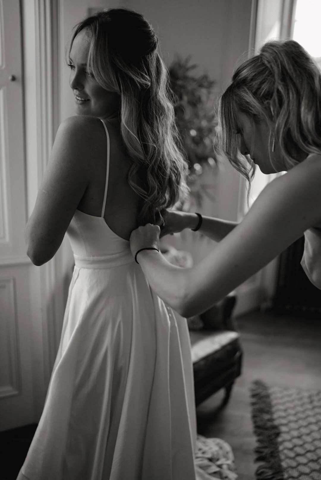 A woman in a white dress is being assisted with her dress by another woman, as she prepares for a special occasion, in a room with a window and a plant in the background.