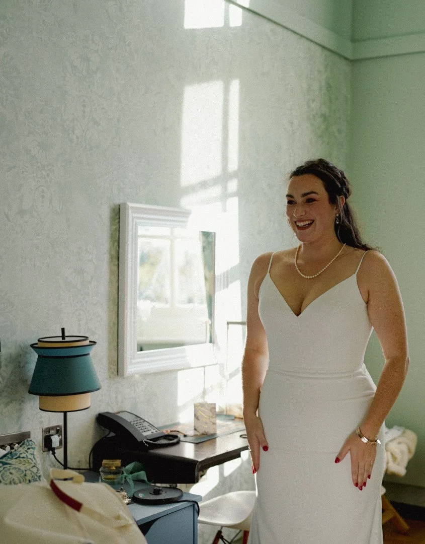 A woman in a white wedding dress stands inside a room with light green walls, smiling happily, with sunlight streaming through a window behind her.