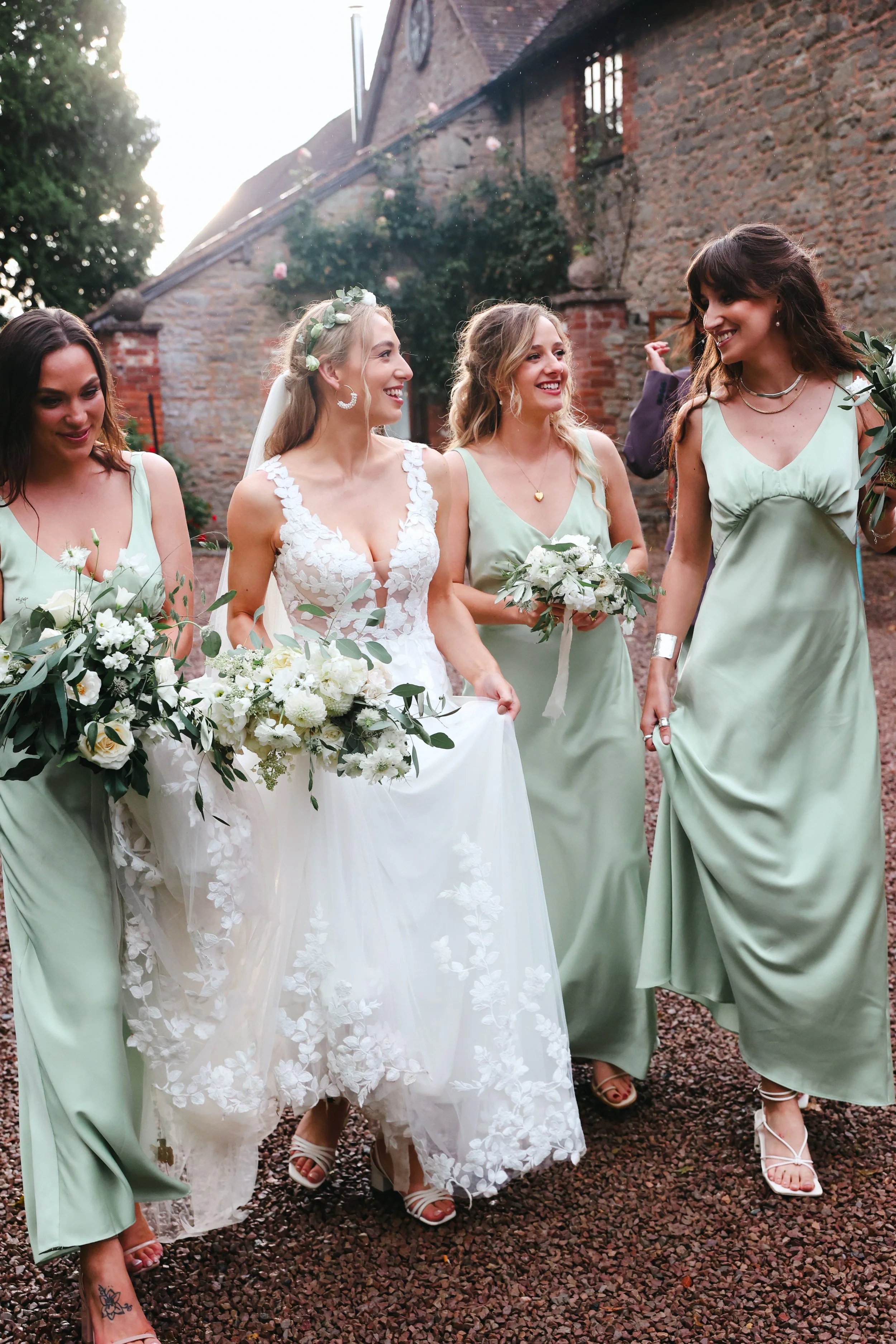A bride and her bridesmaids walking outdoors, smiling, with a rustic brick building in the background. The bride is in a white wedding gown holding a bouquet, and the bridesmaids are in matching light green dresses carrying smaller bouquets.
