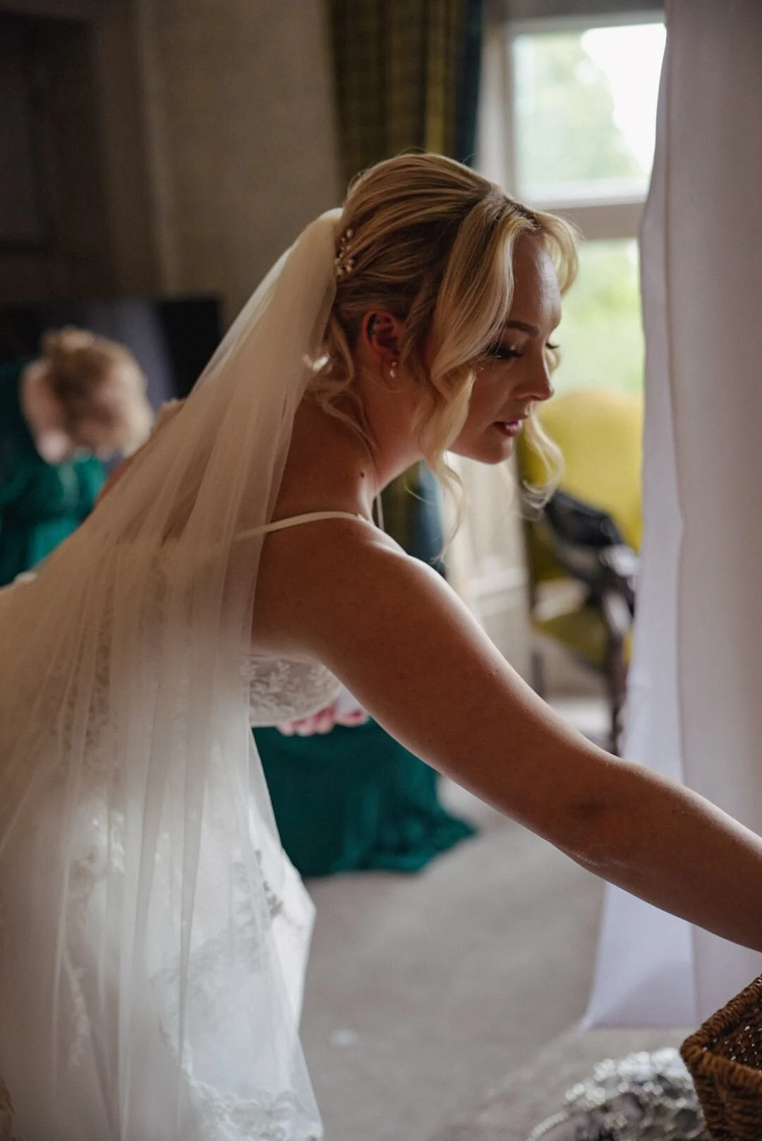A bride with blonde hair styled in loose waves, wearing a veil and a white wedding dress, is standing indoors near a window, looking down and reaching for something.