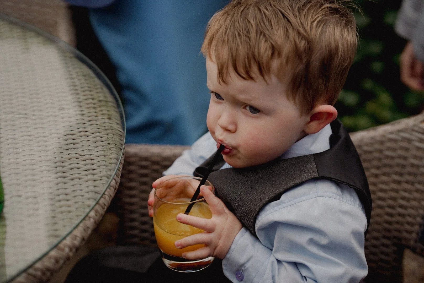 Young boy with red hair drinking orange juice through a straw at a wicker table.