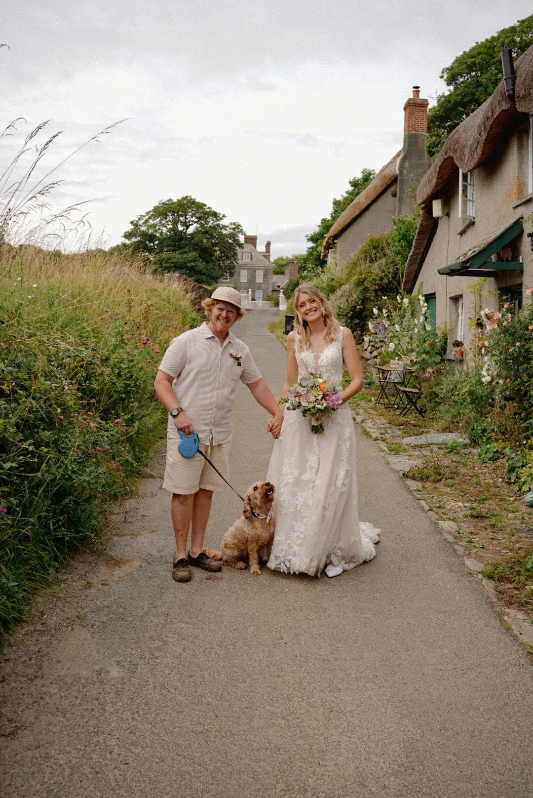 A smiling man and woman in wedding attire holding hands on a rural path, with a brown dog on a leash, greenery, flowering plants, and cottages in the background.