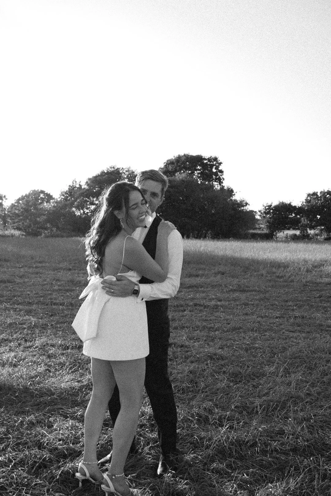 A black and white photo of a smiling woman and man embracing outdoors in a grassy field with trees in the background, during sunset.