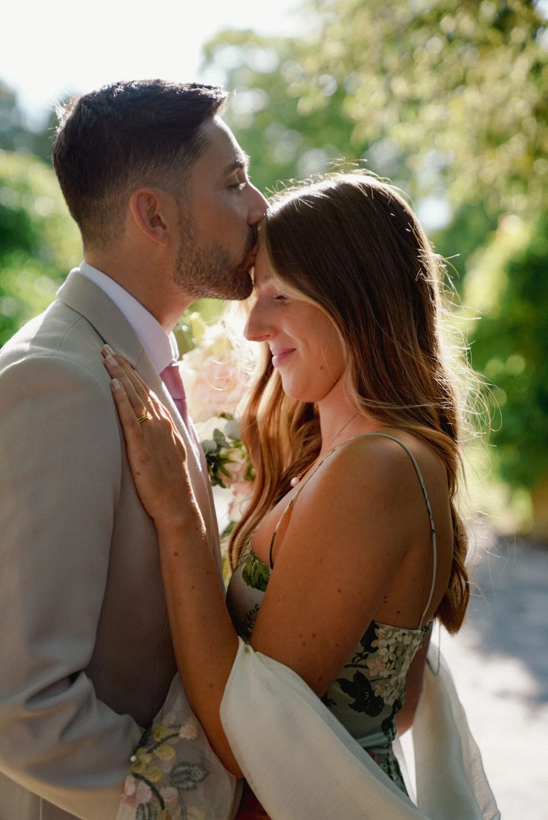 A man and woman share an intimate moment outdoors, with the man gently kissing the woman’s forehead. The woman has long hair and is smiling with her eyes closed, wearing a floral dress, while the man is dressed in a light-colored suit and tie. Green 
