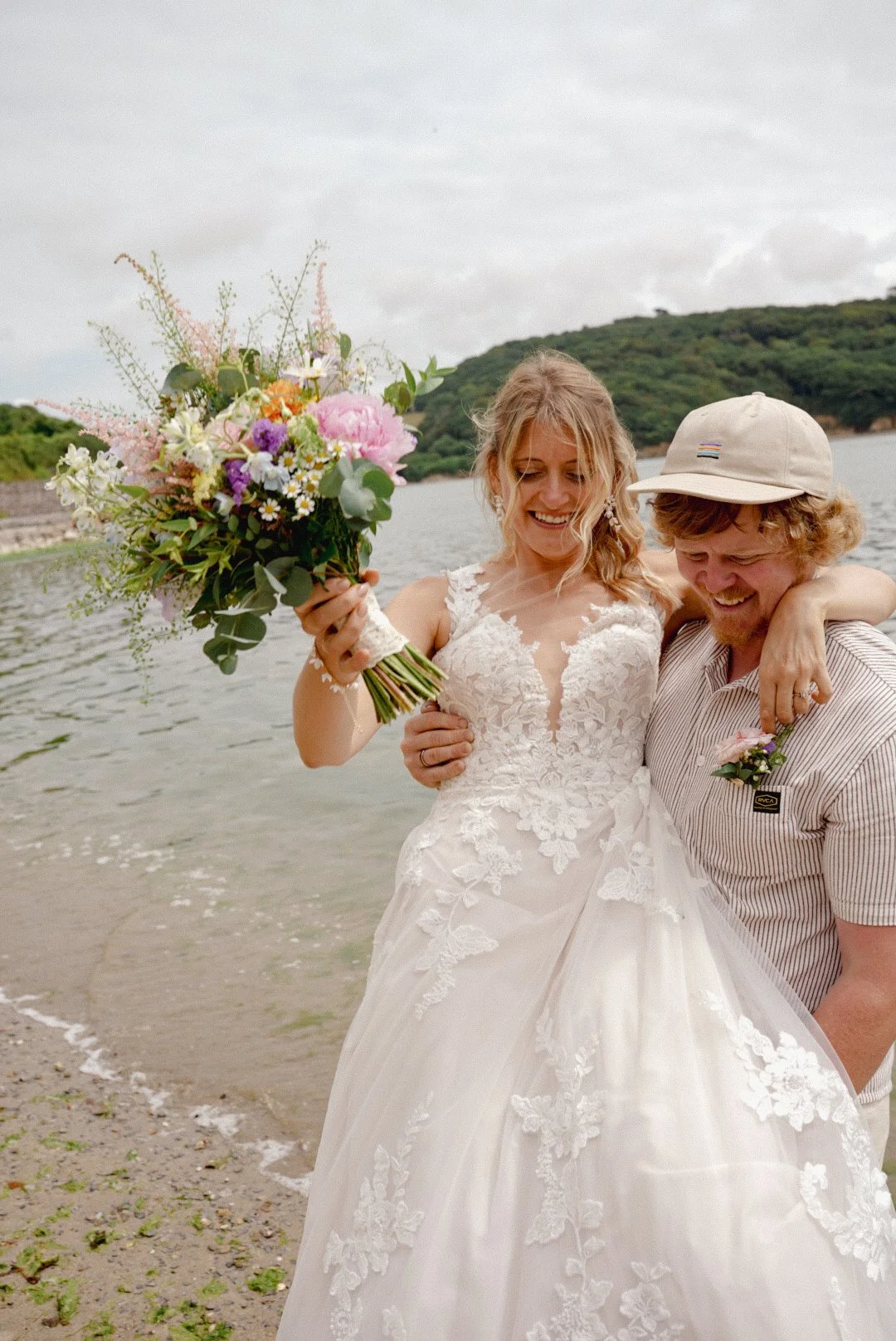 A bride in a lace wedding dress smiling and holding a bouquet of flowers, with a smiling man in casual clothes standing beside her, on a beach with water and hills in the background.
