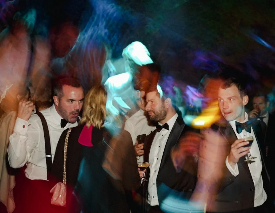 People at a party wearing tuxedos, some holding drinks, with colorful blurred lights and motion effects in the background.
