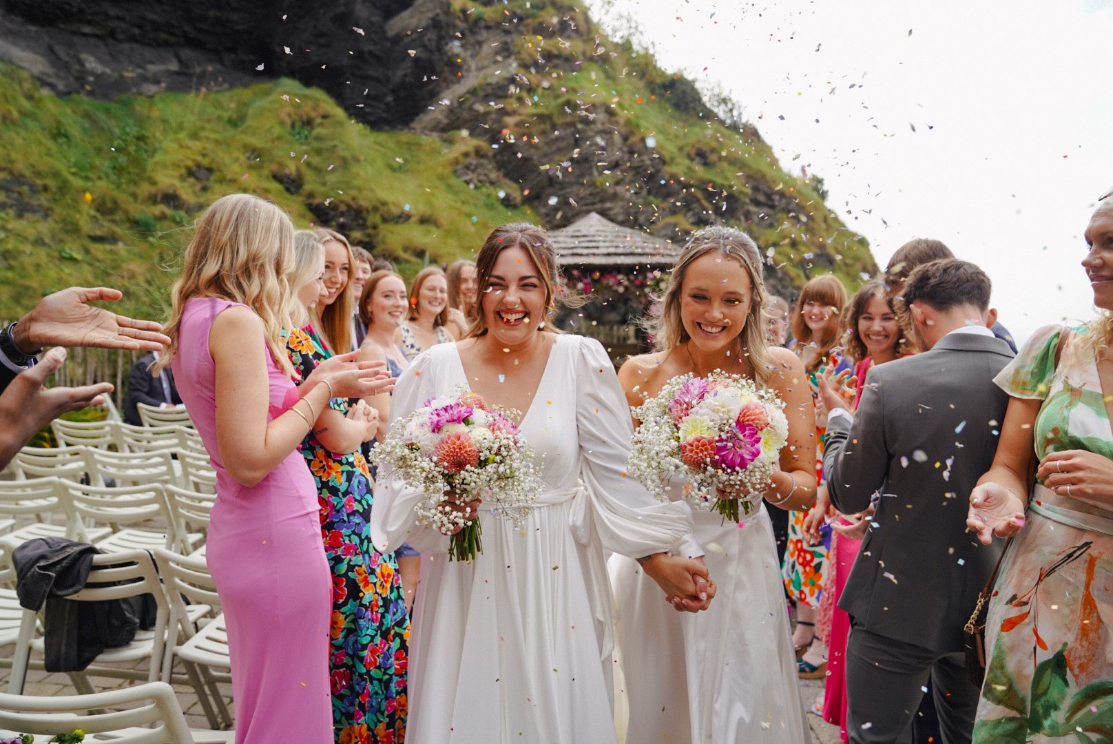 Two brides in white wedding dresses holding hands, smiling, and holding bouquets of flowers, walking through a celebration with friends and family outdoors at a wedding reception.