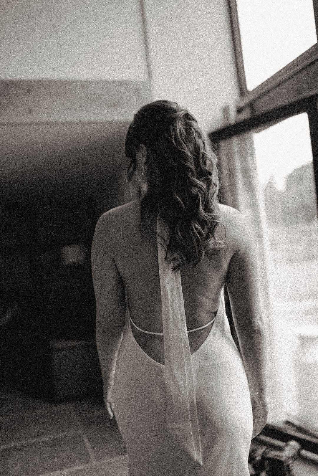 A woman with long curly hair, seen from behind, wearing an elegant backless dress, standing indoors near a large window.