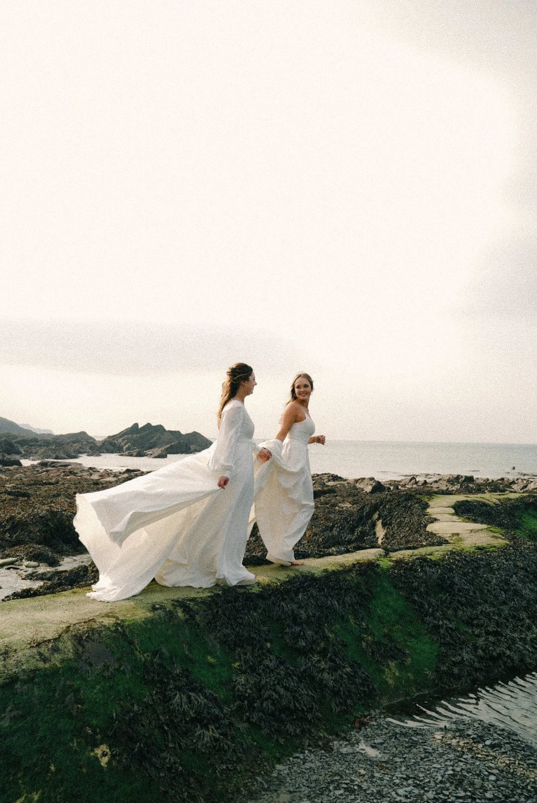 Two women in white dresses walking on rocks near the ocean during sunset.