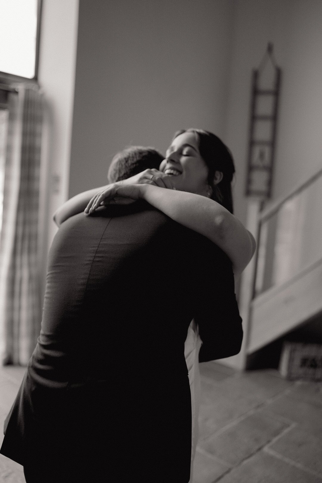A woman with dark hair is hugging a man, smiling and appearing happy indoors near a staircase, in black and white.