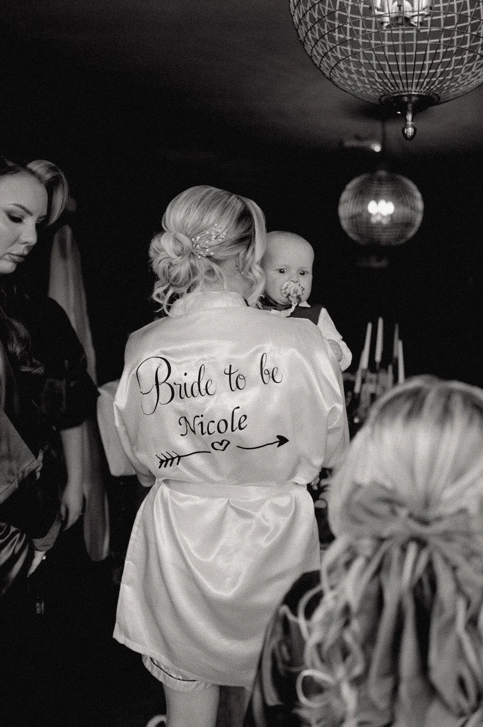 A woman with a satin robe labeled 'Bride to be Nicole' holds a baby at a celebration. The woman has styled hair with a decorative hairpin, and the baby has a pacifier. Guests are nearby, and a large disco ball hangs from the ceiling.