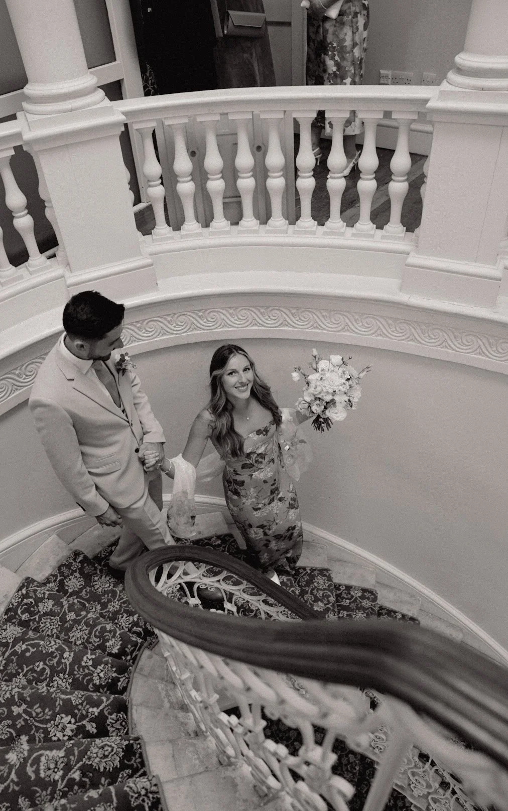 A black-and-white photo of a woman holding a bouquet on a spiral staircase, with a man standing beside her, holding her hand. She is smiling and wearing a floral dress, and he is dressed in a light suit. The staircase has ornate carpet and decorative