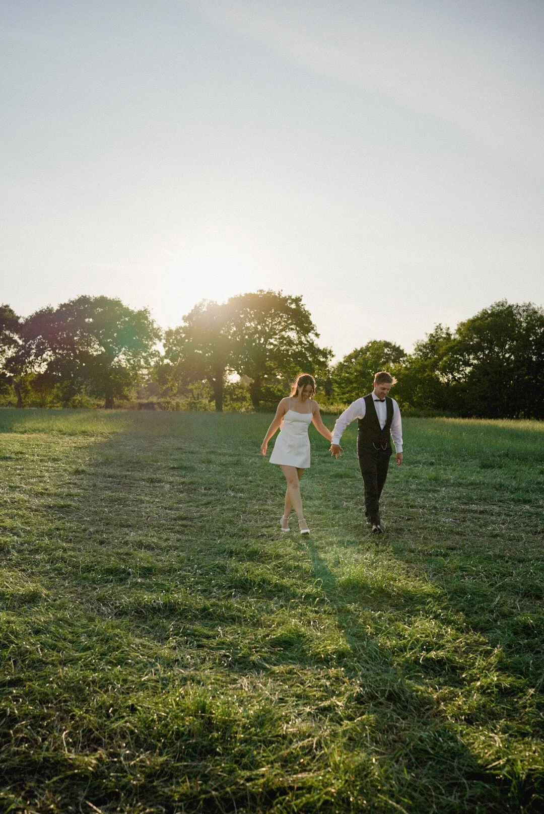 A couple dressed in wedding attire walking hand in hand through a grassy field during sunset.