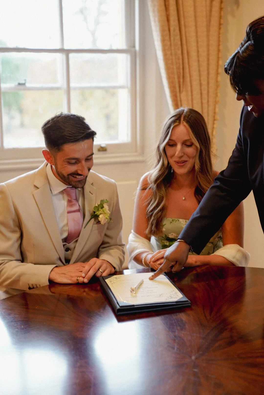 A couple sitting at a table during a wedding ceremony, signing a document with a person pointing at it. The groom wears a beige suit and pink tie, the bride wears a floral dress, and the officiant is in black. There is a window with curtains in the b