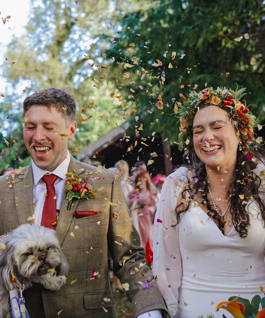 Bride and groom celebrating outdoors with confetti falling, surrounded by friends, with dog in groom's arm, during wedding ceremony.