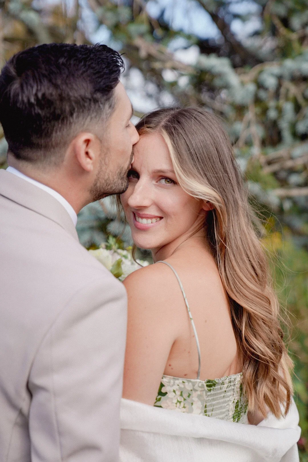 A man kissingly touches his forehead to a smiling woman's cheek as she looks at the camera outdoors, with greenery in the background.