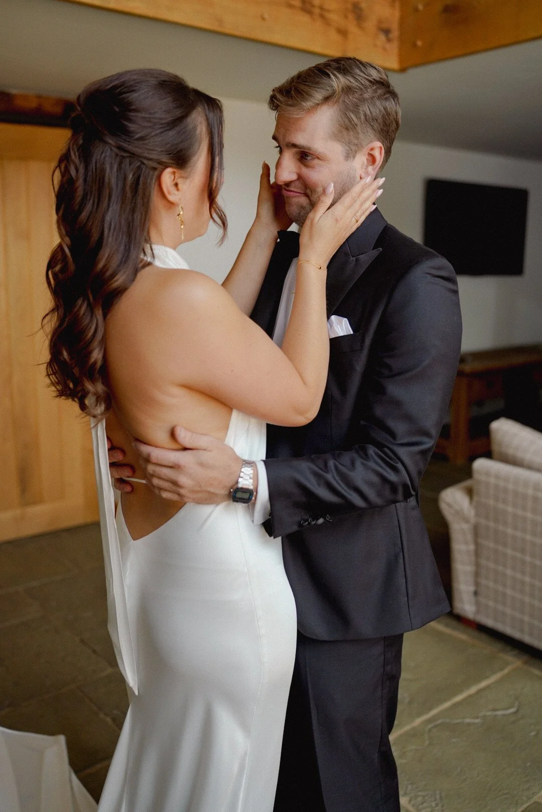 A bride and groom share an intimate moment, with the bride's hands on the groom's face and the groom's arms around her waist, in a warmly lit indoor setting.