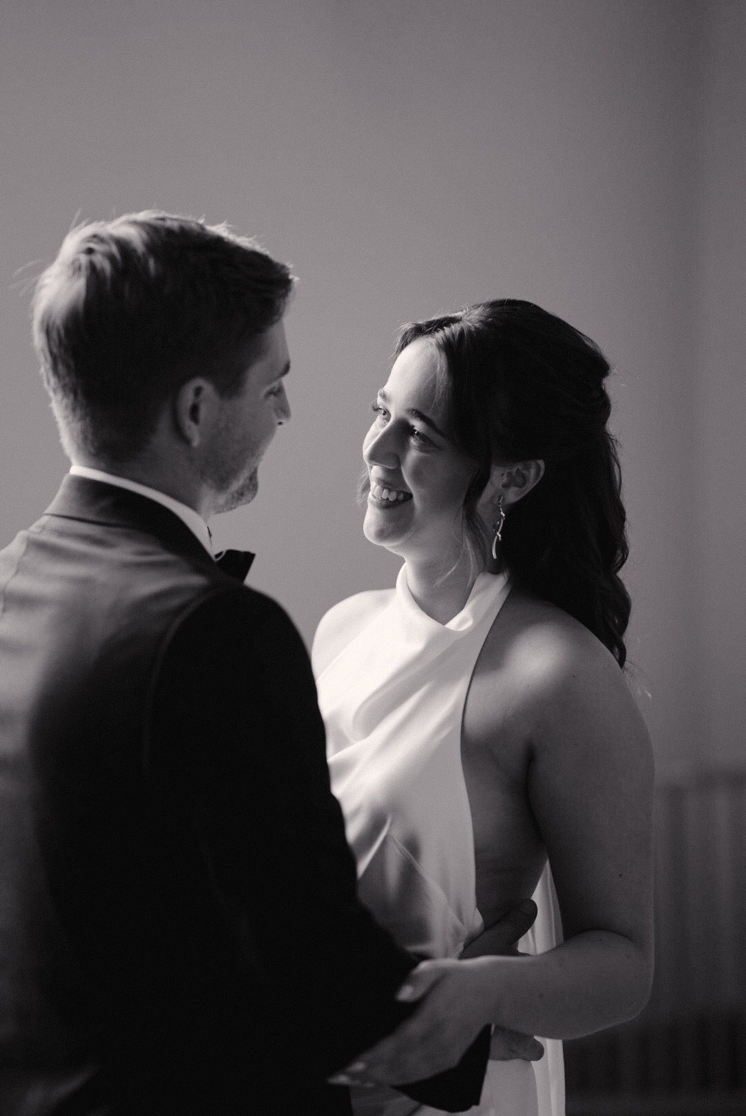 A black and white photo of a happy couple, a man in a tuxedo and a woman in a sleeveless dress, gazing at each other affectionately.