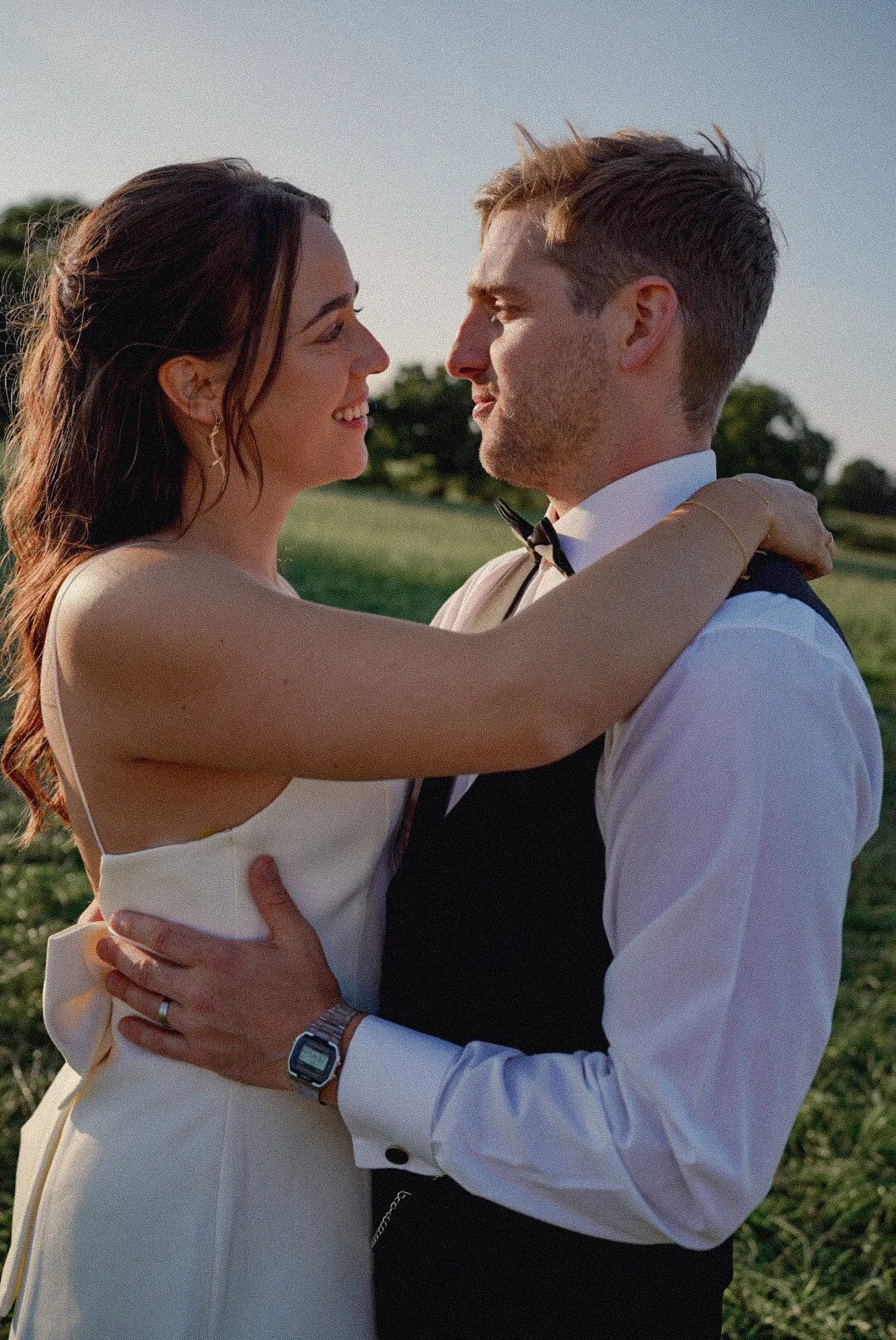 A couple in wedding attire embracing outdoors during sunset, gazing into each other's eyes.