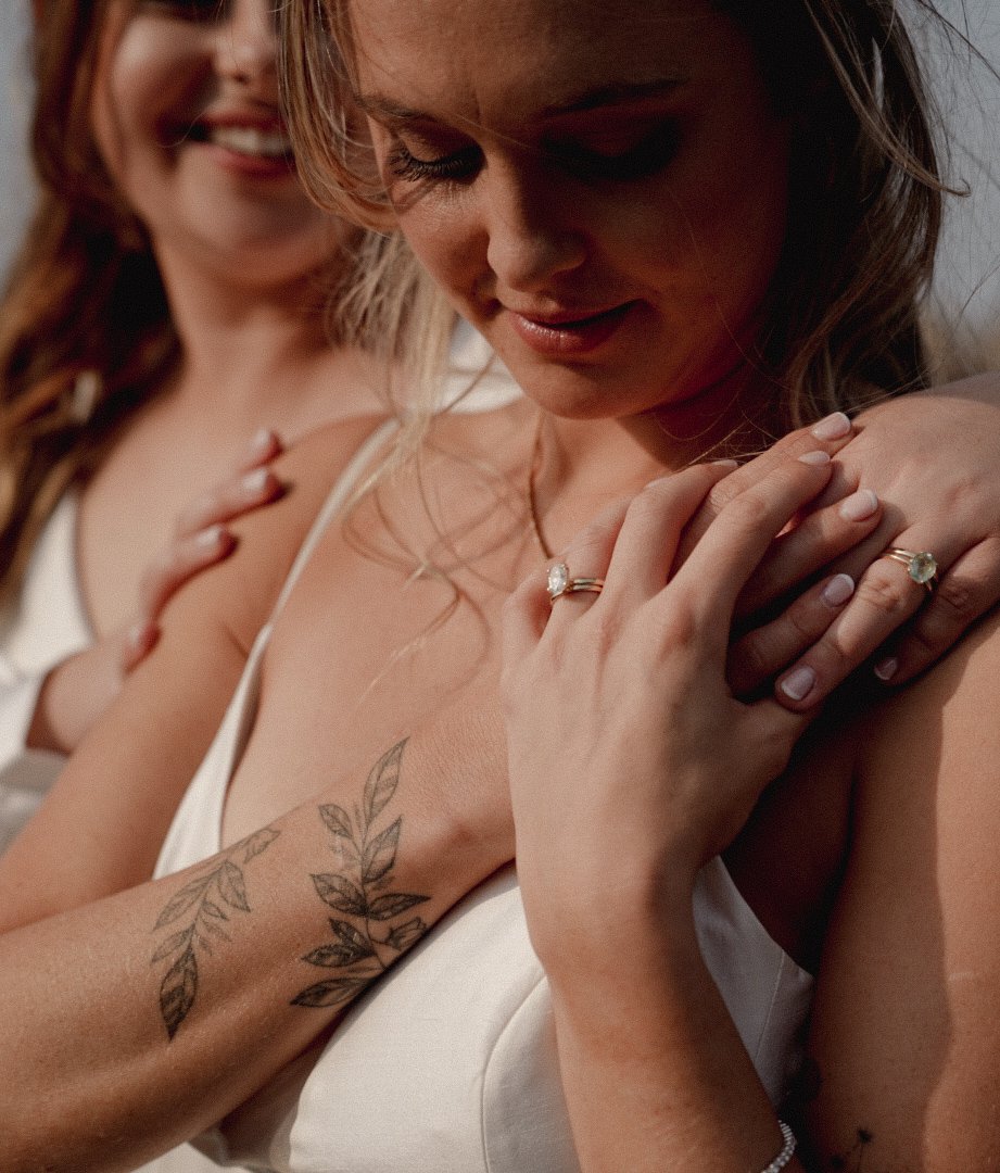 Two women in white dresses, one with a tattoo on her forearm, close together, smiling and touching her shoulder, with rings on their fingers.