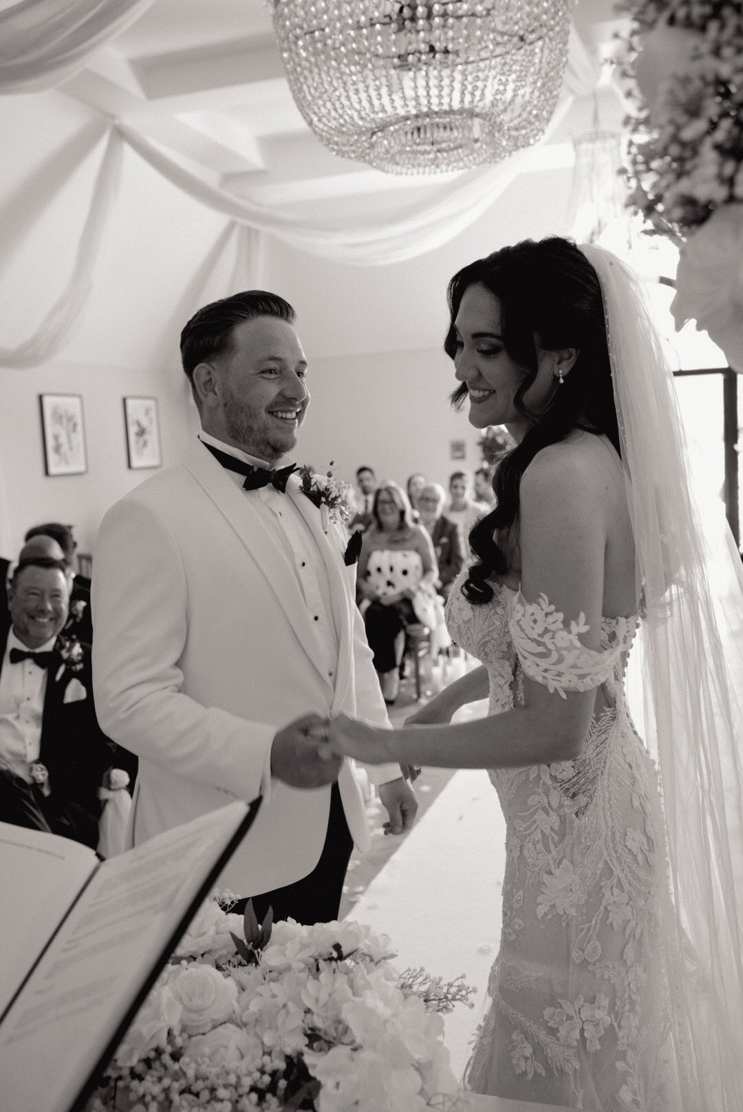 A wedding ceremony with a bride and groom holding hands and smiling, surrounded by seated guests, in a decorated indoor venue.