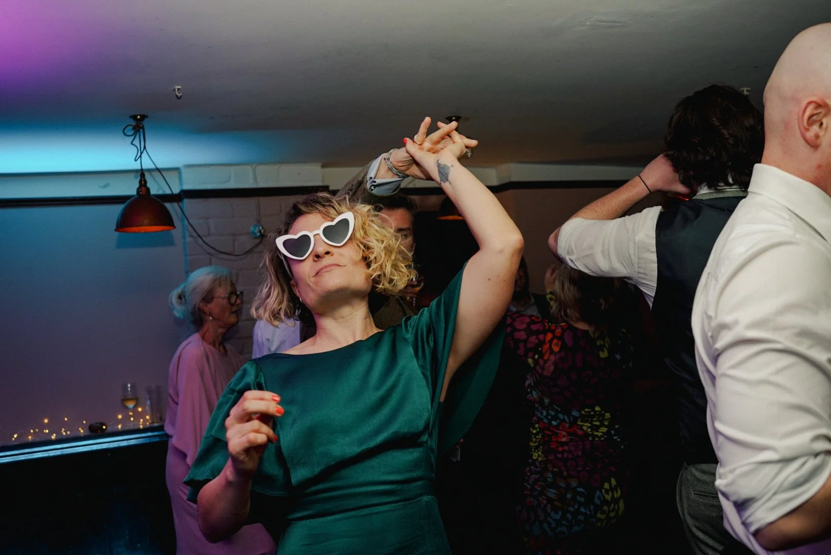 Woman with curly blonde hair wearing large white heart-shaped sunglasses dancing at a party, wearing a dark green dress. Other people are dancing around her in a dimly lit room.