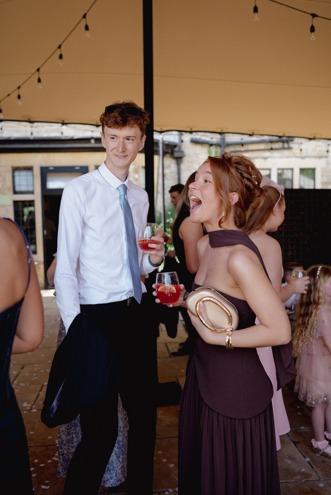 People at a lively party, including a woman in a dark purple dress holding a clutch and talking with a man in a white shirt and tie, both holding drinks. Other guests are visible in the background.