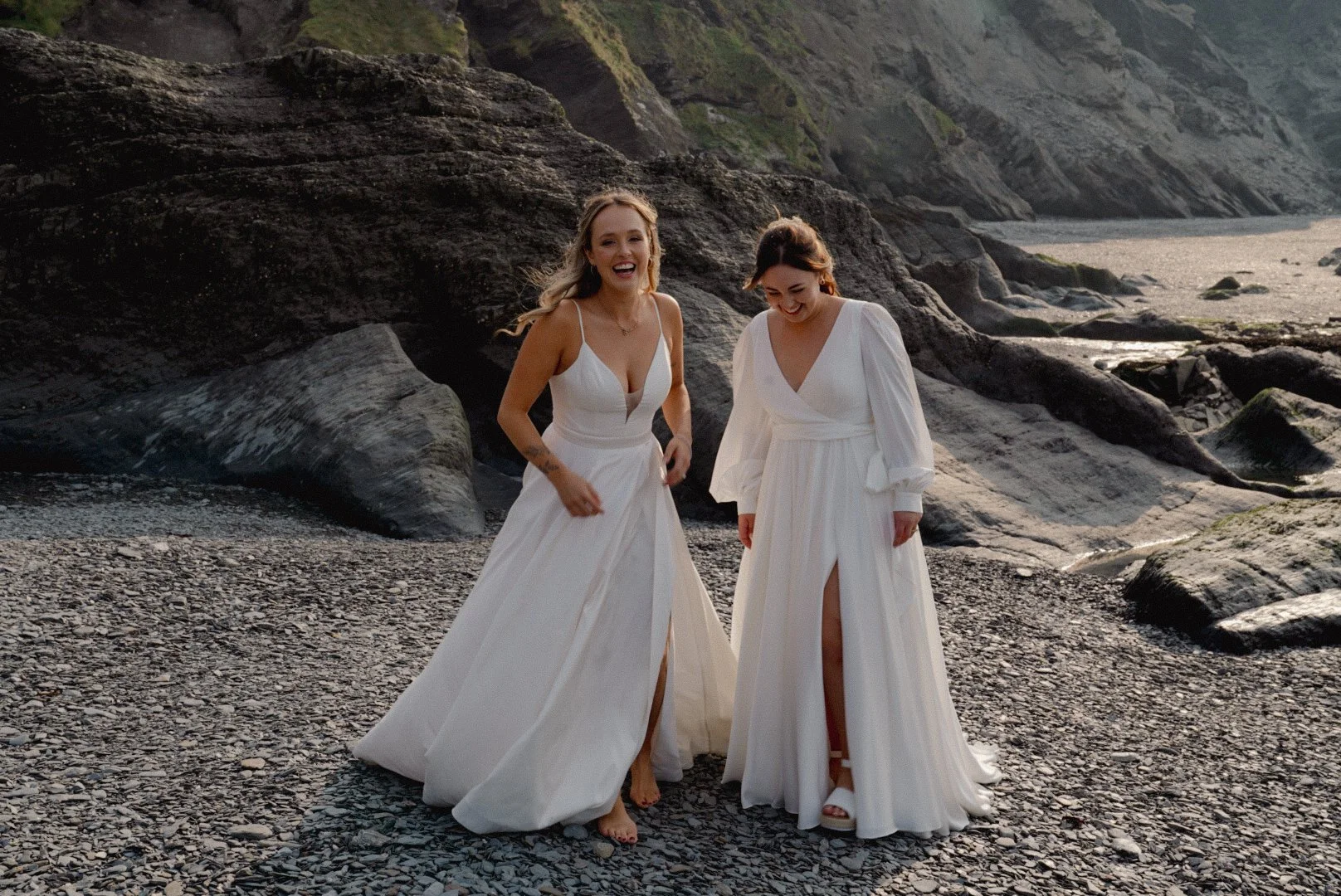 Two women in white dresses walk on a rocky beach, smiling and laughing, with cliffs and the ocean in the background.