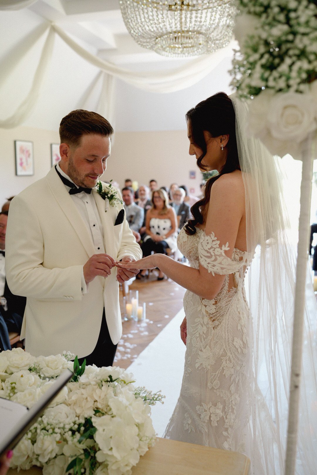 A bride and groom exchange rings during their wedding ceremony, with guests seated in the background.