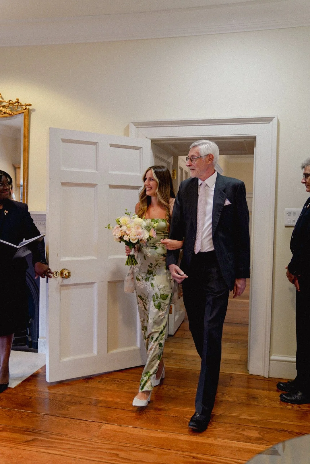 A woman in a floral dress holding a bouquet, walking alongside an older man in a suit, entering a room during a wedding or formal event.
