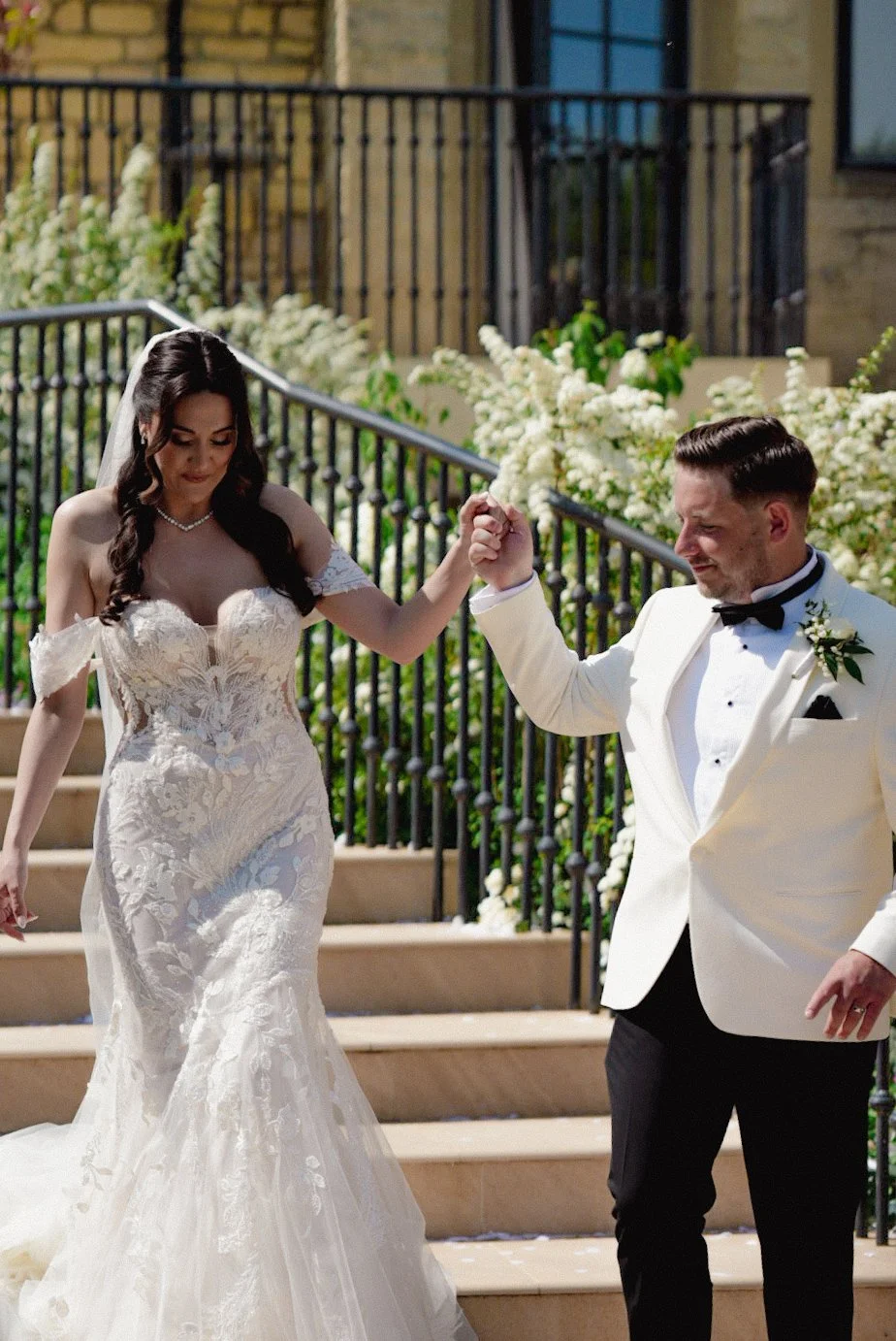A bride and groom holding hands on outdoor wedding stairs, with the bride in a lace wedding gown and the groom in a white tuxedo jacket, black bow tie, and black pants, surrounded by white flowers and greenery.
