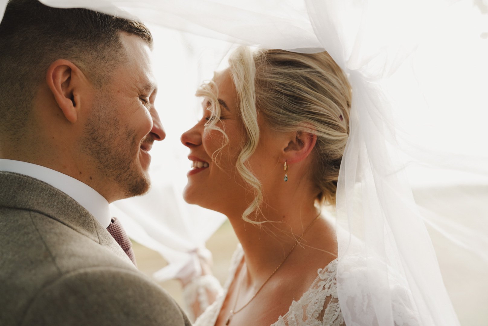 A bride and groom are smiling and about to kiss, their faces close together, with a white veil draped over the bride's head.