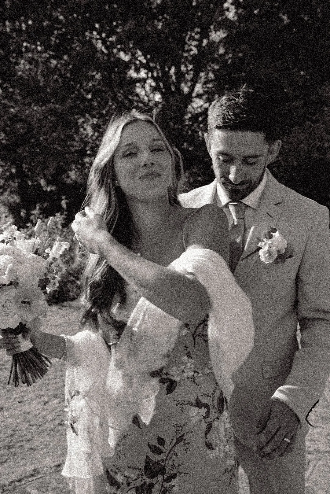 A bride and groom holding hands outdoors, with the bride smiling and the groom looking down, surrounded by trees.