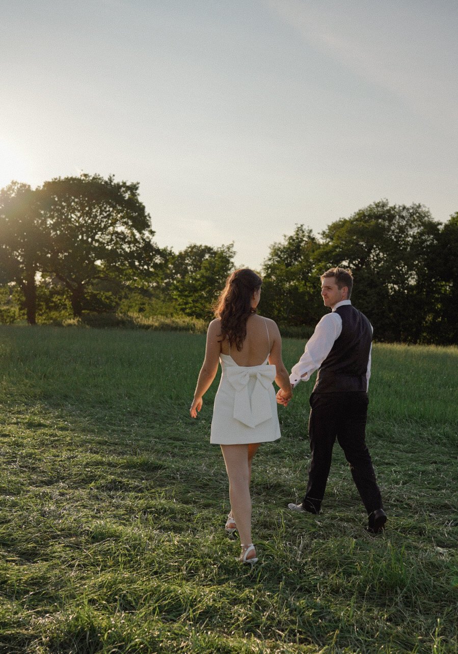 A couple walking hand-in-hand through a grassy field at sunset. The woman is wearing a white dress with a large bow at the back, and the man is dressed in a vest and dress shirt. Trees are visible in the background.