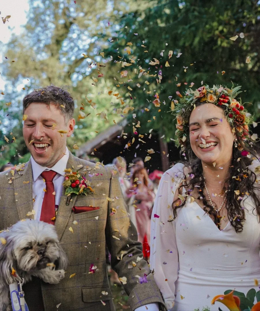 A smiling bride and groom celebrating their wedding outdoors, with colorful confetti falling around them. The groom wears a brown suit with a red tie and boutonniere, and the bride wears a white dress and a floral crown. They are holding hands, and a