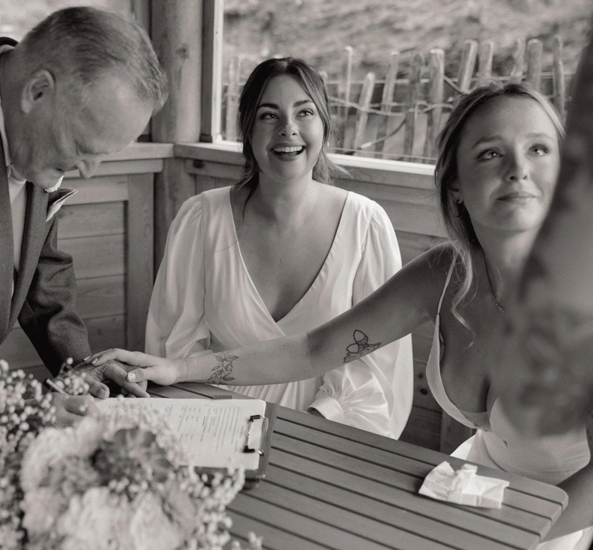 A joyful woman smiling during a wedding ceremony is holding hands with another woman who has tattoos on her arm, as an older man in a suit looks on. They are seated at a table with a bouquet of flowers, inside a rustic wooden building.