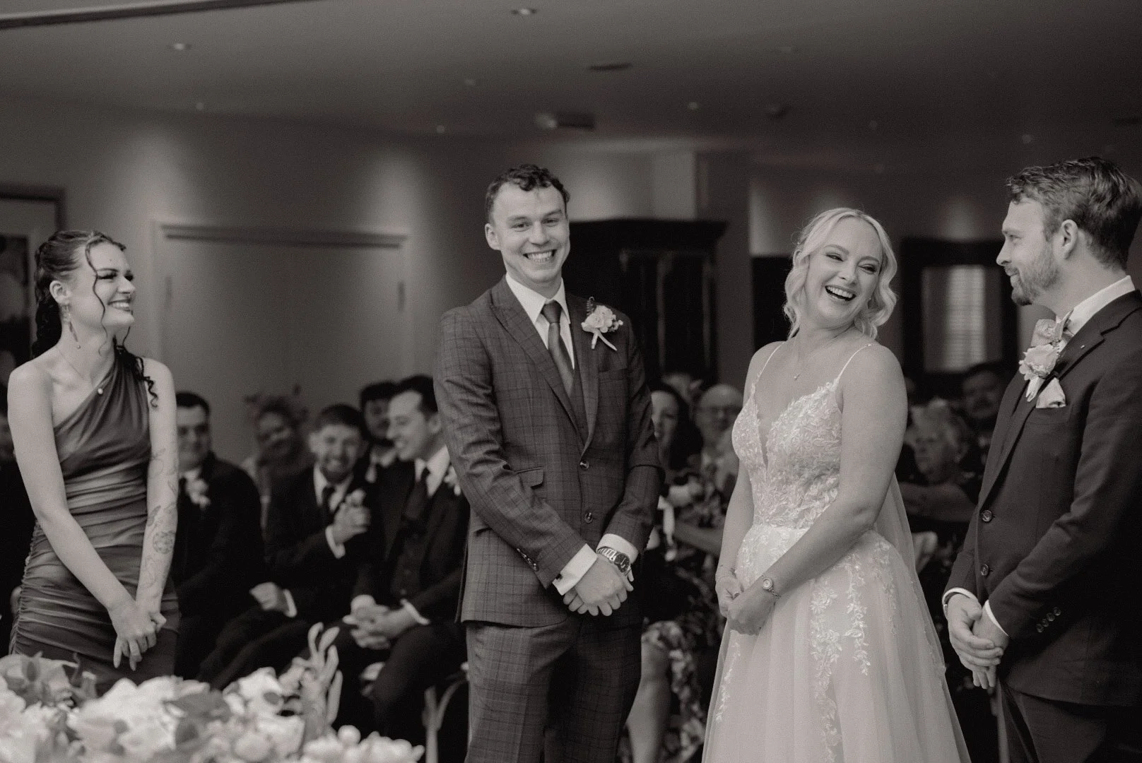 Black and white photo of a wedding ceremony with four people standing in front of seated guests, including the bride and groom smiling and laughing.
