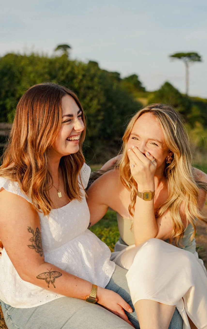 Two women laughing and sitting outdoors in a grassy field with trees in the background during golden hour.