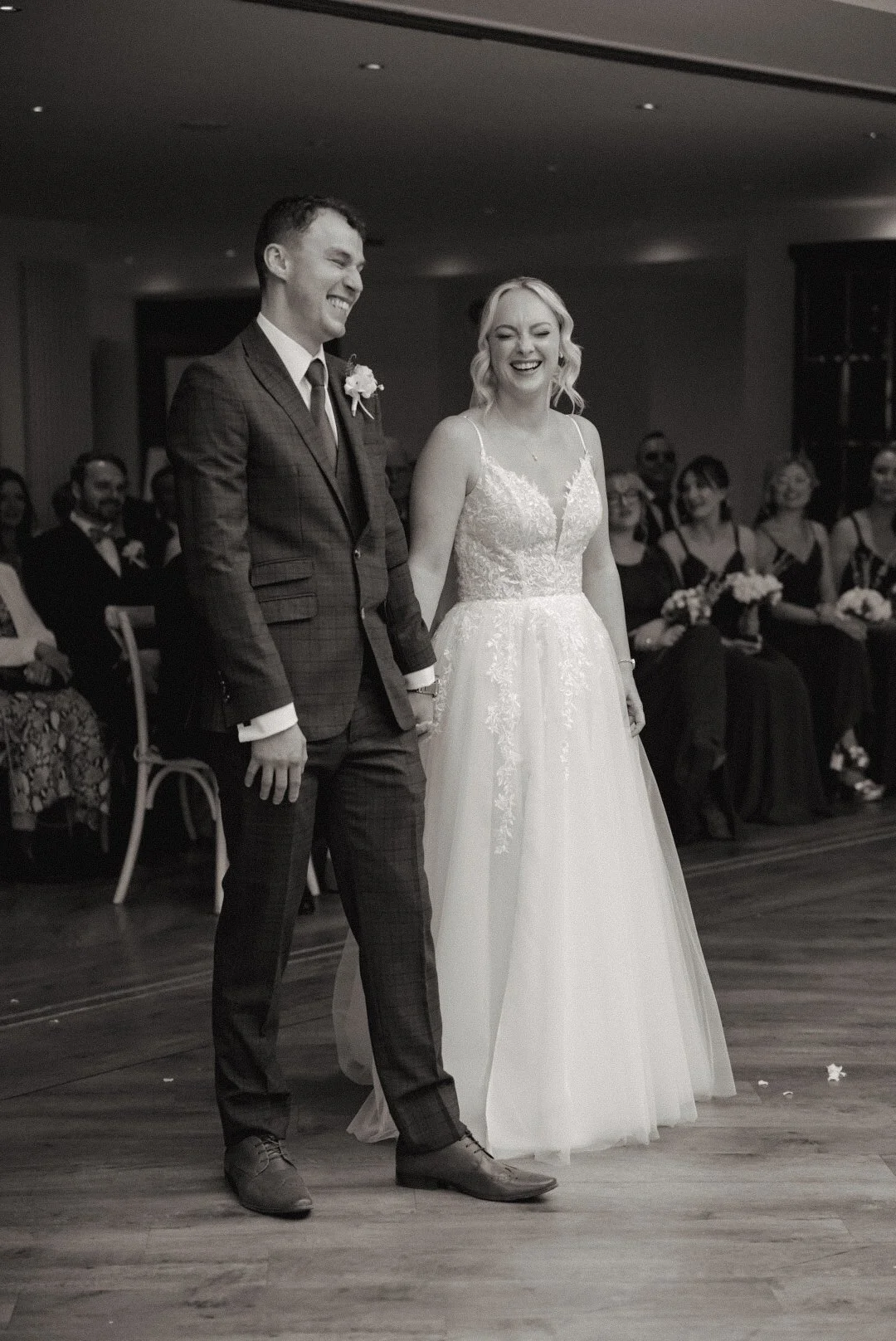 A wedding ceremony with a bride and groom holding hands, smiling, with guests seated in the background.