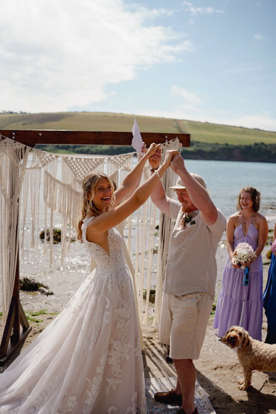 A bride and a man are dancing at a beach wedding, smiling and holding hands, with a woman holding a bouquet and a dog nearby in the background.
