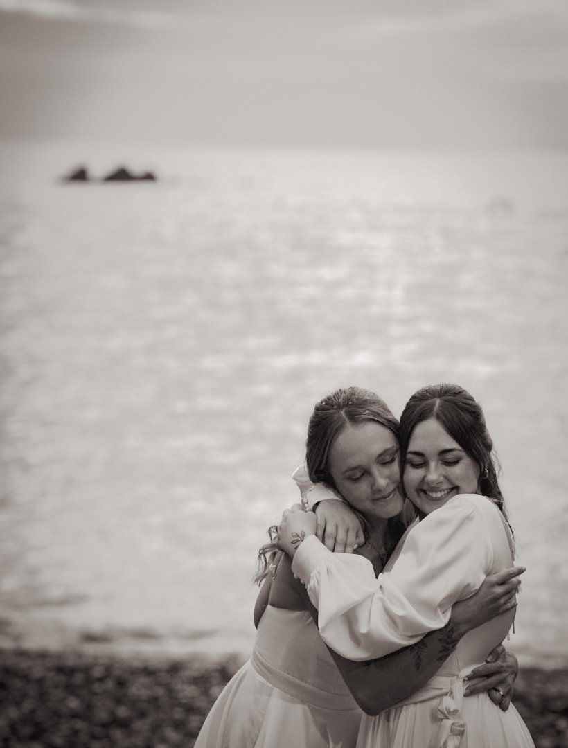 Two women hugging each other happily on a beach with the ocean in the background, black and white photo.