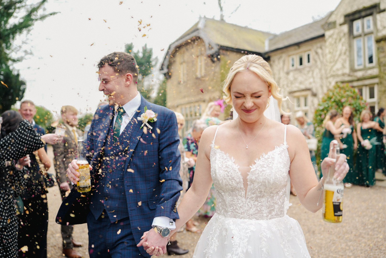 Bride and groom holding hands, smiling with eyes closed, surrounded by guests throwing confetti at outdoor wedding celebration