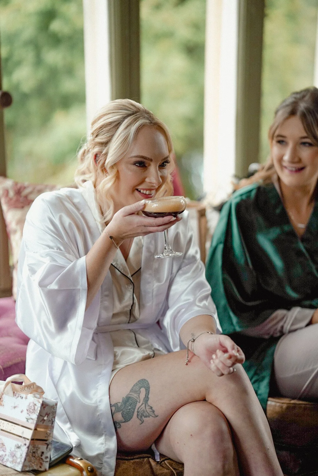 A woman in satin pajamas sitting on a couch, holding a glass of dark beverage, smiling. Another woman in a green robe sits beside her, smiling. The setting appears to be a cozy indoor space with large windows showing greenery outside.