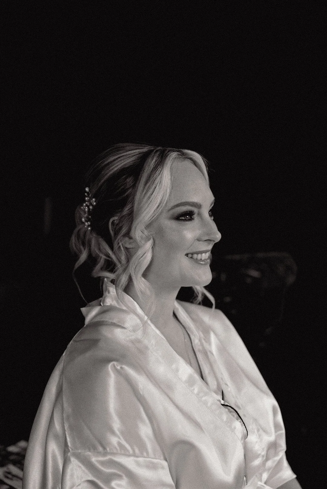 A young woman smiling, with blonde hair styled in loose curls, wearing a shiny satin robe, and a decorative hairpin, in black and white.