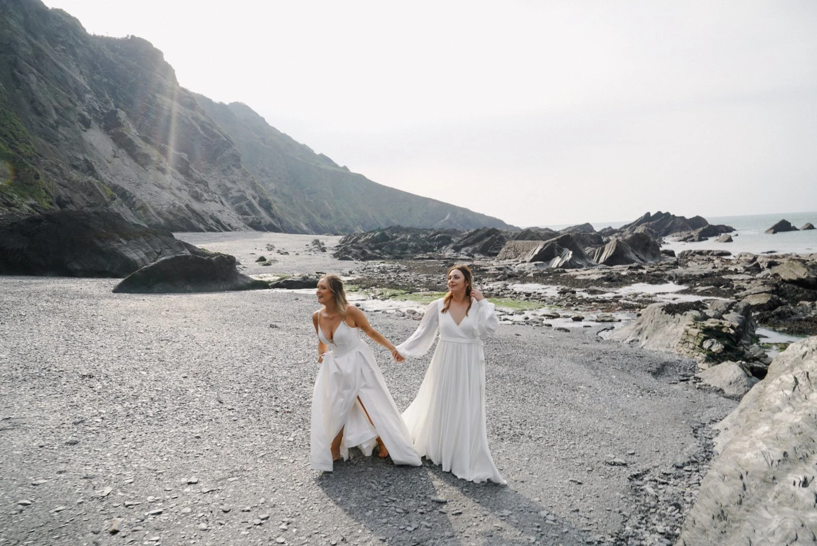 Two women in white dresses holding hands and walking on a rocky beach with mountains and the ocean in the background.