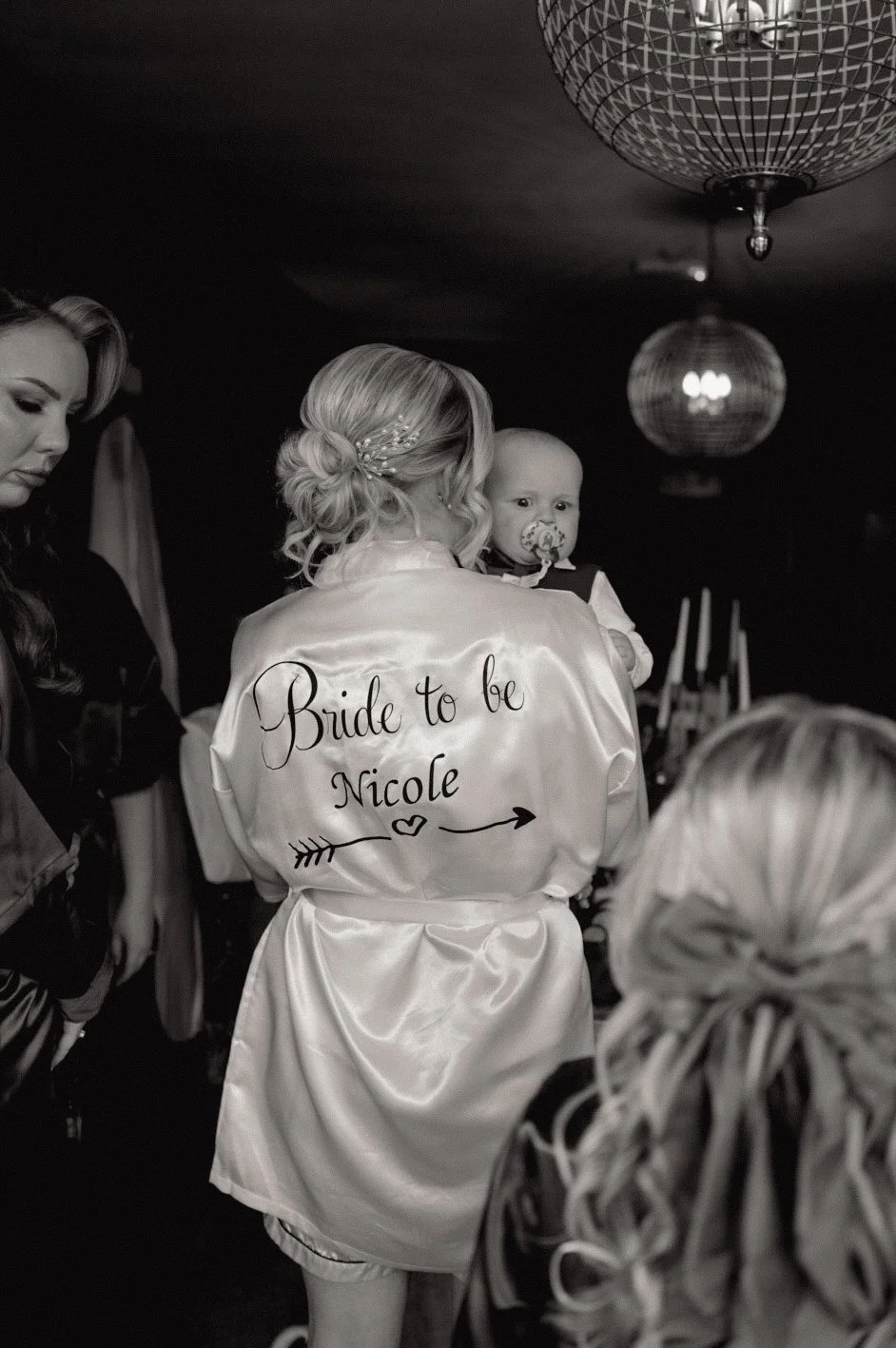 Black and white photo of a bridal party with a woman wearing a satin robe that reads "Bride to be Nicole" on the back, holding a young child with a pacifier, in a dimly lit room with hanging light fixtures.