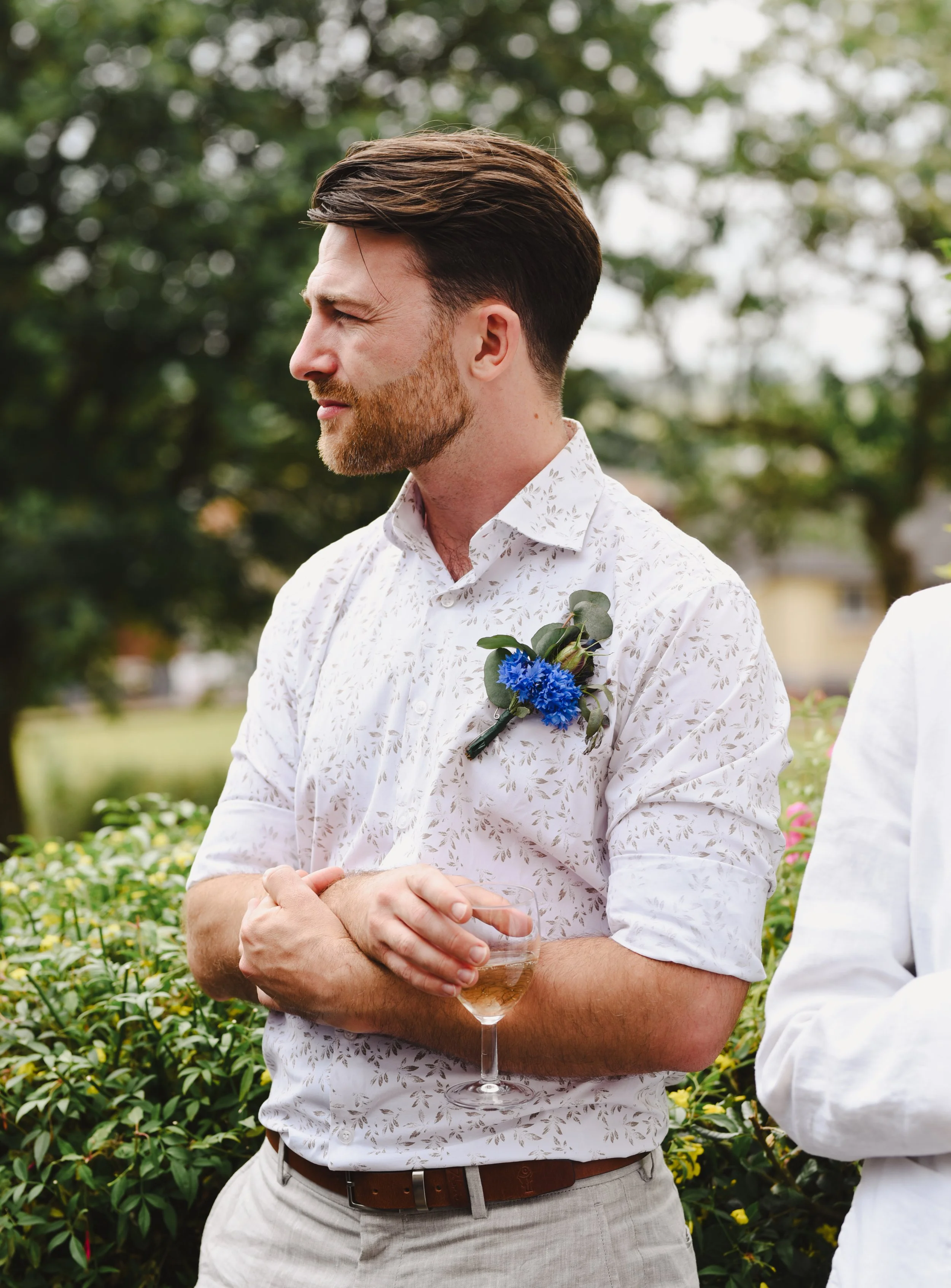 Man with brown hair and beard holding a glass of champagne at an outdoor event, wearing a white floral shirt with a blue boutonniere, standing near greenery.