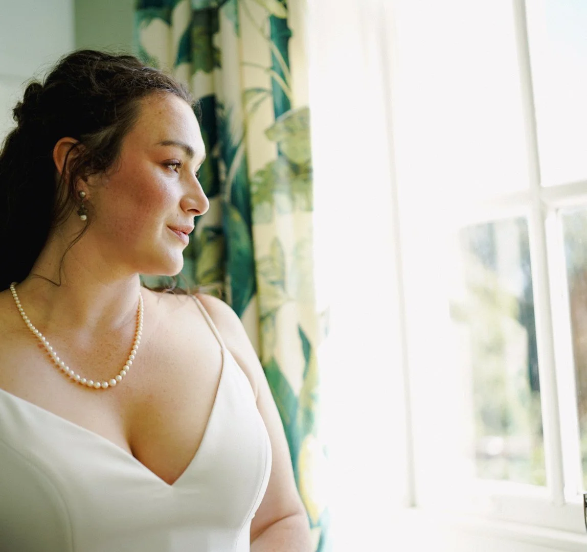 A woman in a white dress with pearl jewelry, standing near a window with floral curtains, looking outside thoughtfully.