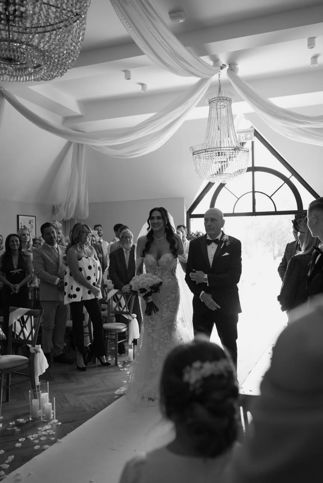 Bride walking down the aisle with her father during a wedding ceremony, surrounded by guests in a decorated indoor venue with a chandelier and draped fabric.